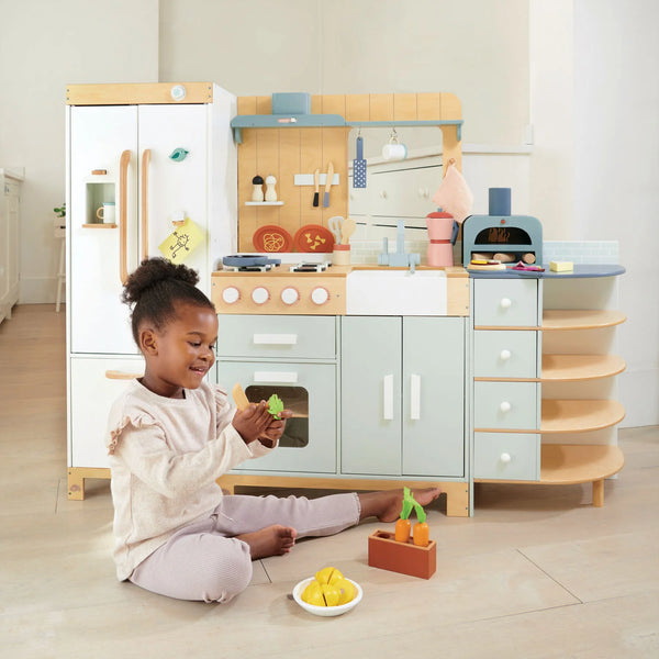 A young girl sits on the floor playing with toy fruits in front of the La Fiamma Grand Kitchen, a kids kitchen set featuring pastel colors, a wooden play kitchen design, and various kitchen accessories.