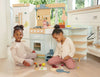 Two young children sit on the floor playing with toy kitchen items and pretend food. Behind them is the La Fiamma Grand Kitchen—a pastel-colored wooden play kitchen set featuring a charming pizza oven toy—inside a bright, sunlit room.