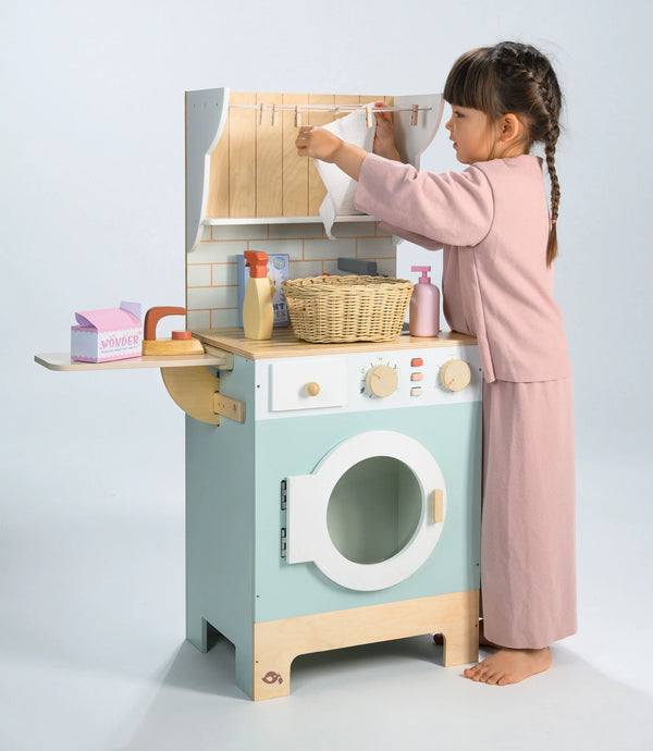 A young child in pink pajamas plays with the Wooden Laundrette, a charming wooden laundry toy washing machine. She hangs a cloth on the mini clothesline, using themed accessories like the included basket and pretend soap—inviting little ones into creative pretend play laundry fun.