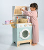 A young child in pink pajamas plays with the Wooden Laundrette, a charming wooden laundry toy washing machine. She hangs a cloth on the mini clothesline, using themed accessories like the included basket and pretend soap—inviting little ones into creative pretend play laundry fun.