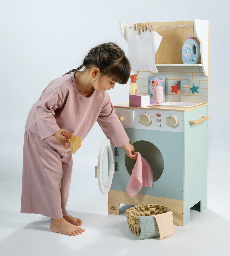 A young child in pink pajamas engages in pretend play, placing a small cloth into the Wooden Laundrette toy washing machine, with a basket and folded fabric from the laundry play set nearby.