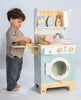 A young child stands beside the Wooden Laundrette, pretending to iron clothes. This laundry play set includes detergent bottles, a basket, and towels, encouraging imaginative play and teaching household chores.