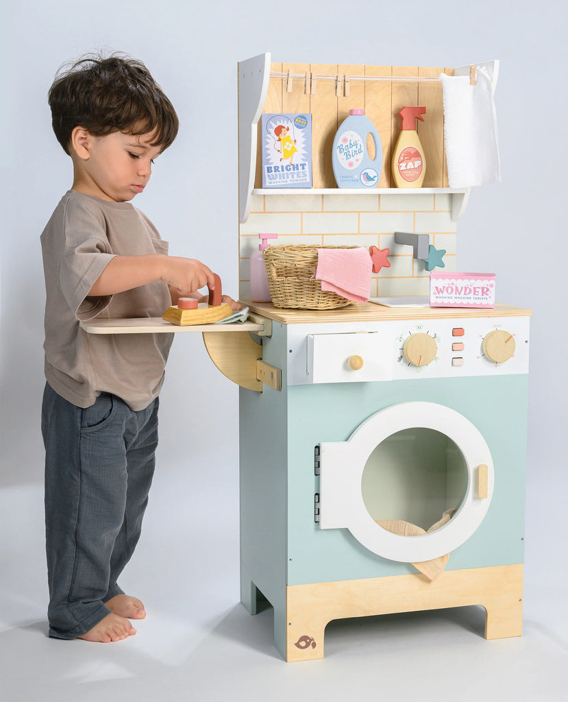 A young child stands beside the Wooden Laundrette, pretending to iron clothes. This laundry play set includes detergent bottles, a basket, and towels, encouraging imaginative play and teaching household chores.