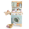 Wooden Laundrette playset with towels, detergent bottles, soap, and a basket. Perfect for imaginative laundry adventures, this wooden laundry toy features shelves above that hold cleaning supplies and a hanging towel; the washing machine's round door is open to reveal a towel inside.