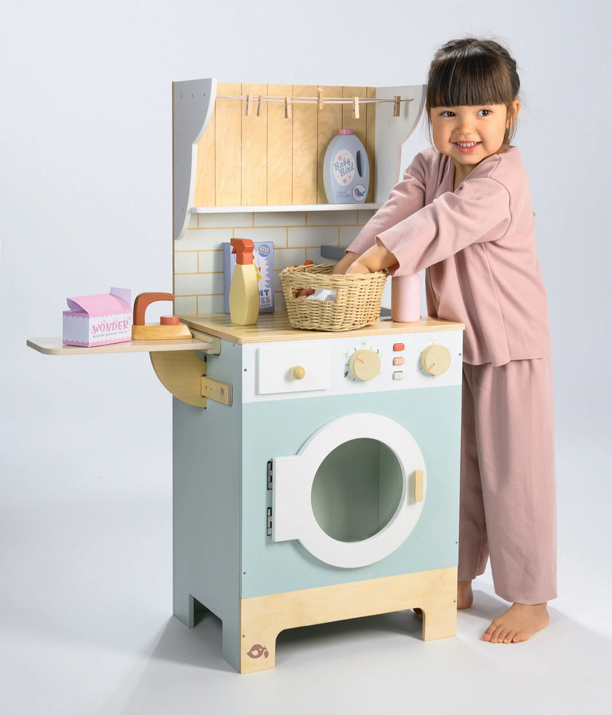 A young child in pink pajamas smiles while engaging in pretend play laundry, joyfully playing with the Wooden Laundrette set, pretending to do laundry with a wicker basket and toy detergent bottles.