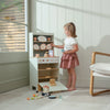 A young girl plays with the Kitchen Dresser and Dishwasher, pretending to cook and arrange toy dishes. She stands on tiled flooring near a window with sunlight streaming in, and a wooden chair is nearby. The dishwasher unit sits beside her kitchen dresser, enhancing the fun of her imaginative play.
