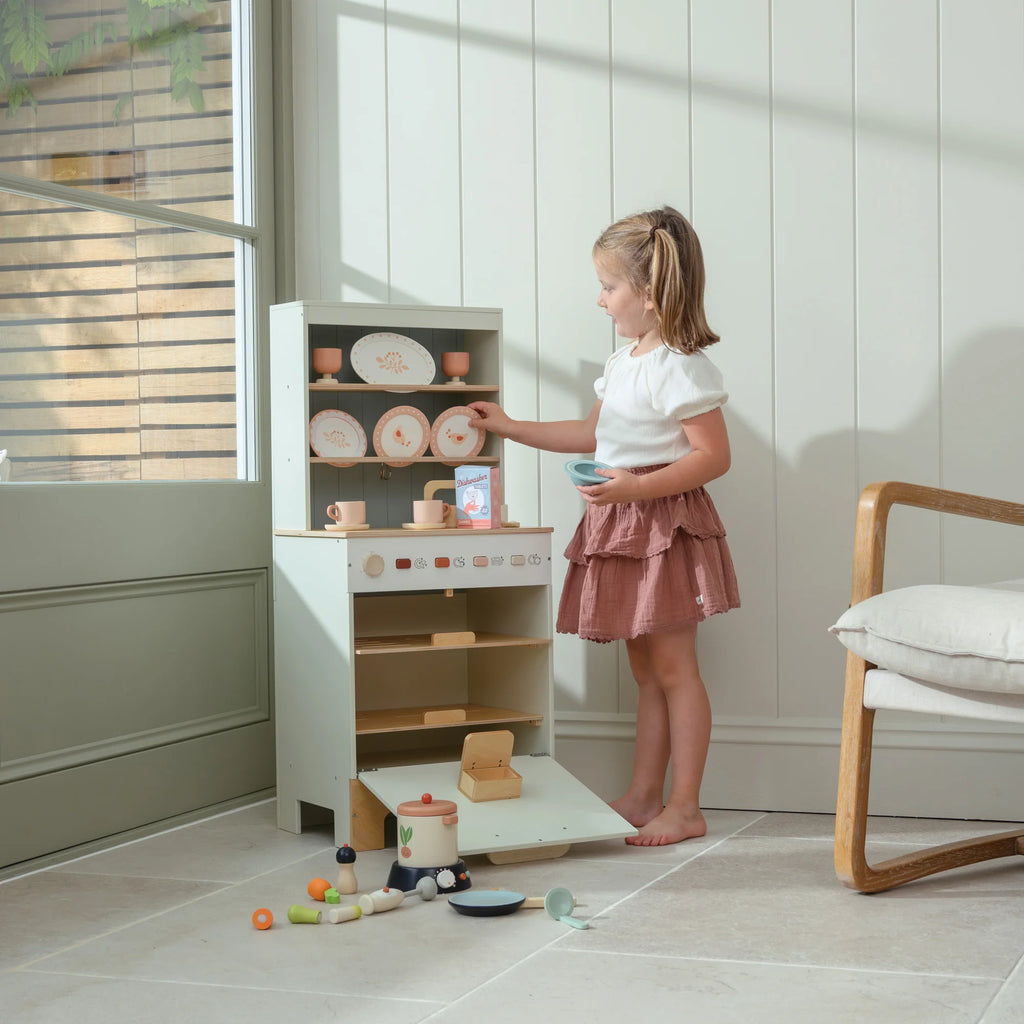 A young girl plays with the Kitchen Dresser and Dishwasher, pretending to cook and arrange toy dishes. She stands on tiled flooring near a window with sunlight streaming in, and a wooden chair is nearby. The dishwasher unit sits beside her kitchen dresser, enhancing the fun of her imaginative play.