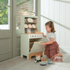 A young girl in a white shirt and pink skirt plays with the Kitchen Dresser and Dishwasher, arranging toy dishes and pretend food on the floor beside her in a sunlit room. The realistic Kitchen Dresser and Dishwasher add extra charm to her imaginative play.