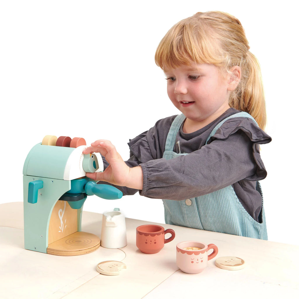 A young girl in overalls plays barista at her pretend café, placing a cup under the Babyccino Maker’s spout. Toy cups, cookies, and a milk jug are arranged on the table in front of her against a white background.