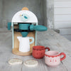 The Babyccino Maker features a wooden coffee toy with a bird design, two cups with faces, a white milk jug, and two cookies—ideal for pretend café play.