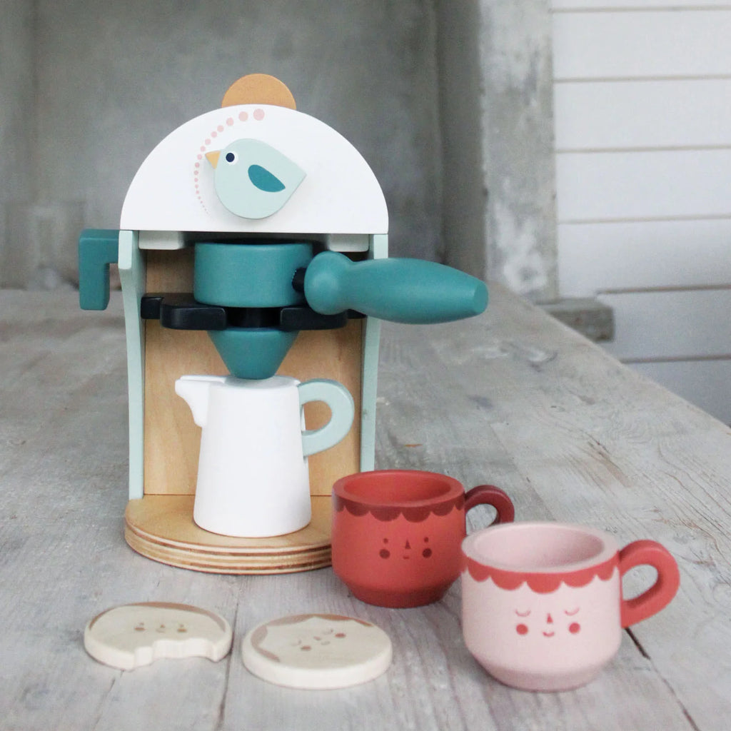 The Babyccino Maker features a wooden coffee toy with a bird design, two cups with faces, a white milk jug, and two cookies—ideal for pretend café play.