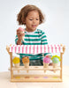 A young child with curly hair is playing with an Ice Cream Scoops and Smiles toy, holding a toy ice cream cone, while smiling and looking at the colorful ice cream toys.