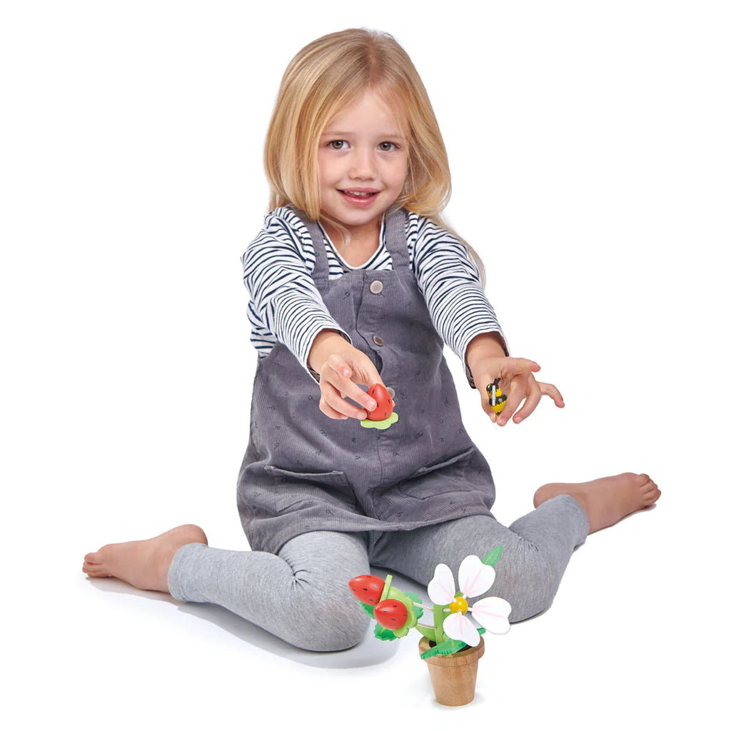 A young girl with blonde hair sits cross-legged on a white background, playing with colorful magnetic Strawberry Flower Pot toys, wearing a striped top and gray overalls.