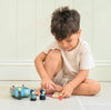 A young boy sits on the floor, smiling and playing with the Badger Picnic Motorbike Ride toy and small animal figurines, dressed in a light outfit against a pale wall.