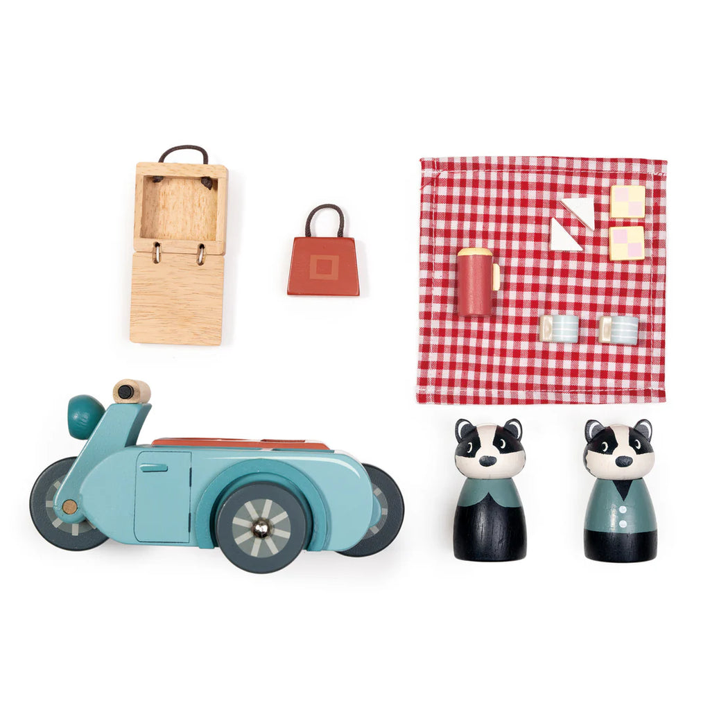 The Badger Picnic Motorbike Ride playset includes a blue toy motorbike, two wooden badger figures, a wooden suitcase, red handbag, and a checkered picnic blanket with tiny plates and cups—ideal for countryside fun.