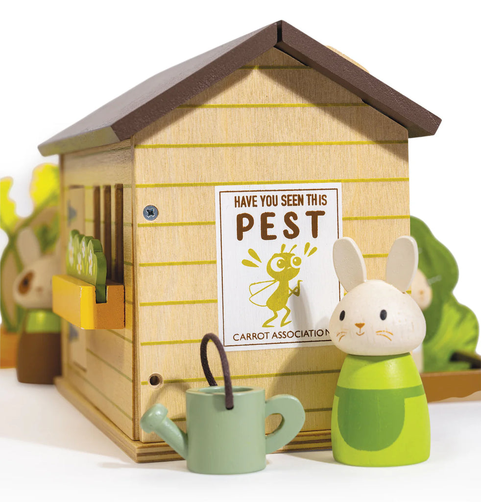 Standing by the charming wooden dollhouse—adorned with a whimsical “Have you seen this pest?” poster featuring a cartoon bug—the Bunny’s Carrot Patch playset showcases its adorable wooden bunny alongside toy vegetables and a green watering can, creating a playful garden scene.