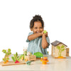 A smiling child in a green checkered dress plays with Bunny’s Carrot Patch, surrounded by wooden toy vegetables, a bunny rabbit toy, a tea set, and a small wooden dollhouse on a white background.