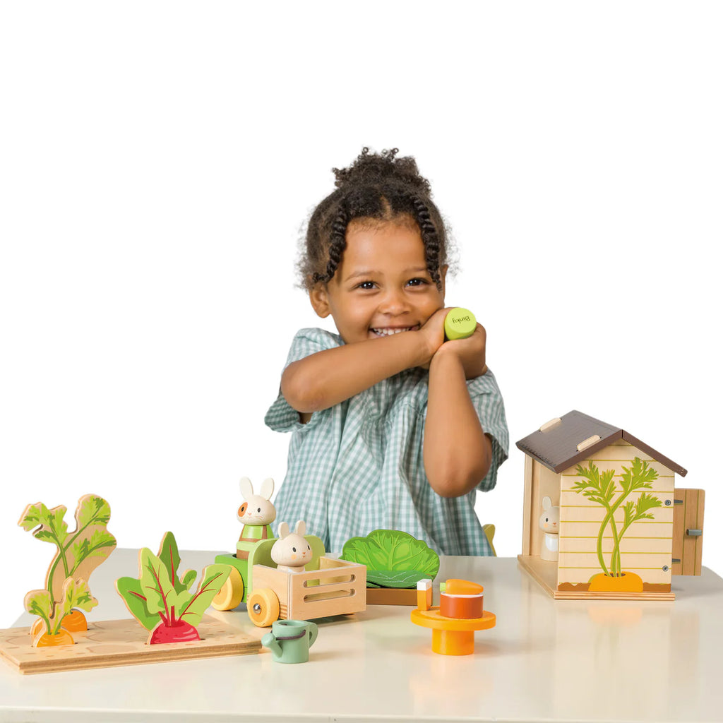 A smiling child in a green checkered dress plays with Bunny’s Carrot Patch, surrounded by wooden toy vegetables, a bunny rabbit toy, a tea set, and a small wooden dollhouse on a white background.