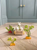 Three bunny rabbit toys sit in the Bunny’s Carrot Patch—a charming wooden cart surrounded by a vegetable garden playset, featuring wooden toy plants shaped like vegetables—all placed on a light wood floor with blue cabinets in the background: perfect for enhancing any wooden dollhouse scene.