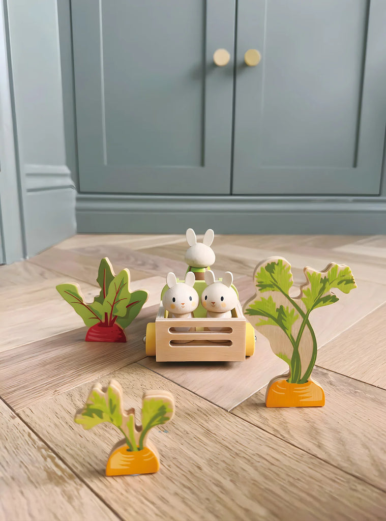 Three bunny rabbit toys sit in the Bunny’s Carrot Patch—a charming wooden cart surrounded by a vegetable garden playset, featuring wooden toy plants shaped like vegetables—all placed on a light wood floor with blue cabinets in the background: perfect for enhancing any wooden dollhouse scene.