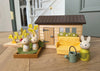 The Bunny’s Carrot Patch set features delightful bunny rabbit toys, a yellow sofa, garden plants, a watering can, and a green door, all charmingly arranged on a wooden floor indoors.
