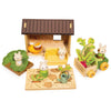 Bunny’s Carrot Patch is a wooden toy set that includes bunny figures, a charming wooden dollhouse, a vegetable garden playset with a crate of crops on a cart, a watering can, and other garden-themed accessories arranged for imaginative play.