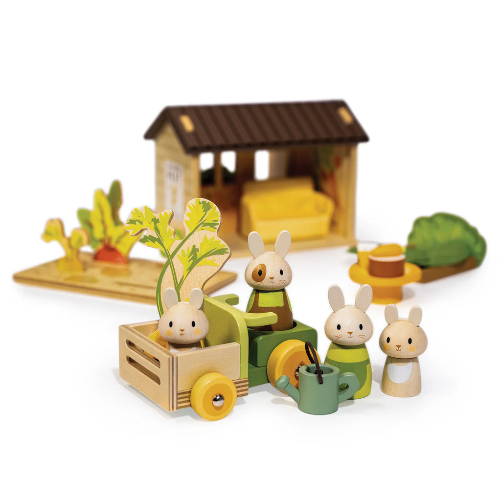 Four wooden bunny rabbit toys from Bunny’s Carrot Patch sit near a green tractor, cart, watering can, and a yellow hat, with a barn, vegetables, and a charming vegetable garden playset scene in the background.