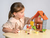 A young girl plays with Robin's Nest Box, a small wooden dollhouse with an orange roof, open sides, and toy accessories—including a bird family—arranging figures and furniture on the table.