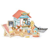 A colorful wooden beach shop playset titled "Sandy's Beach Hut" featuring toy figures and accessories like a deck chair, fish and chips, a wooden toy seagull, and a boat