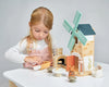 A young girl engaged in imaginative play with a Penny Windmill toy set including a windmill, arches, and figurines on a white table, focused and assembling pieces with care.