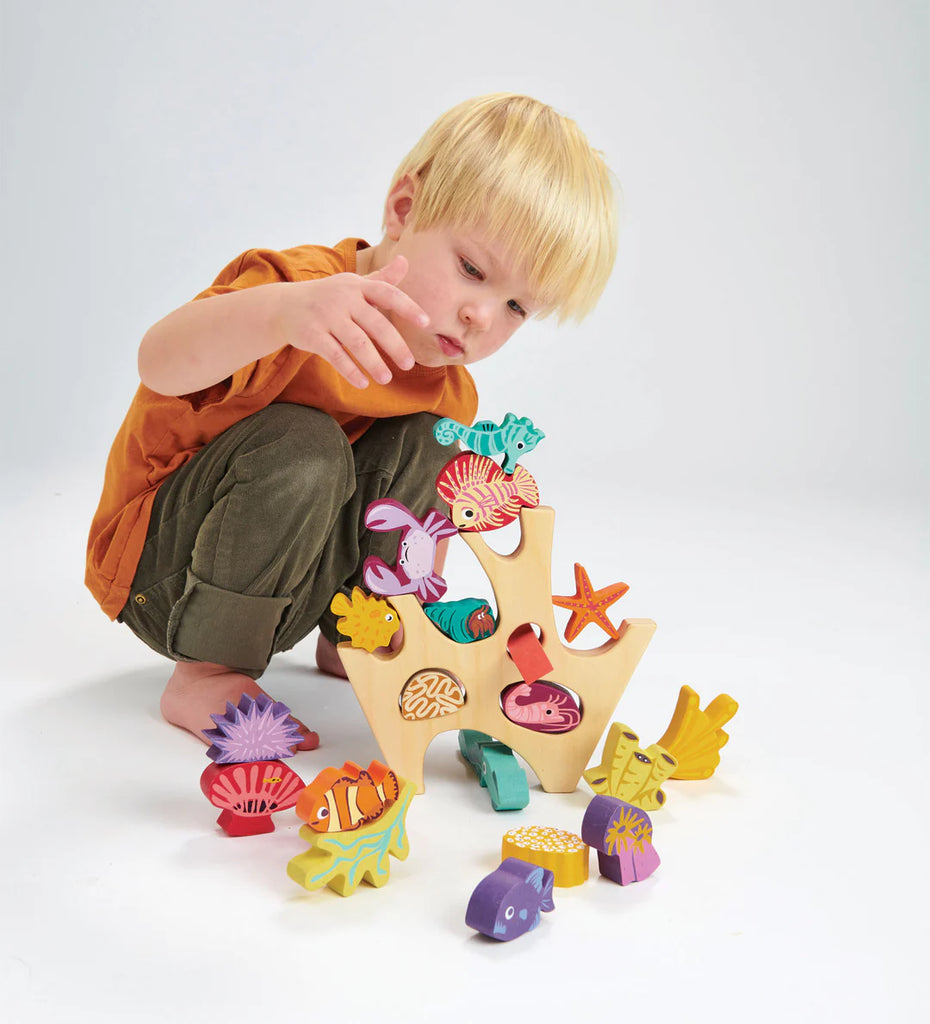 A young child with blond hair plays with a Stacking Coral Reef set, carefully arranging the colorful wooden sea creatures and boat on a white background.