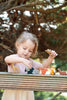 A young girl, suitable for the age range of 3 years and older, is playing with the Woodland Animals Set on a wooden surface outdoors, surrounded by greenery. She is holding a giraffe.