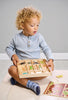 A young child with curly hair sits on the floor, looking surprised while holding a Bug Hotel wooden puzzle box with garden bugs shapes. He wears a light blue shirt and denim shoes.