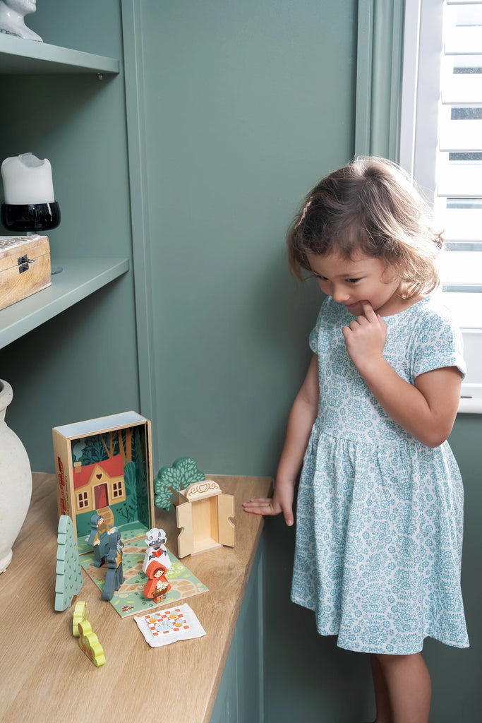 A young girl in a light blue patterned dress stands by a wooden table, gazing thoughtfully at "Your Story of Little Red Riding Hood," a woodland-themed display of colorful wooden toy figures and buildings.