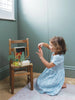A young girl in a blue patterned dress kneels on a rug, playing with the "Your Story of Little Red Riding Hood" set arranged on a wooden chair in a softly lit room with pale green walls.