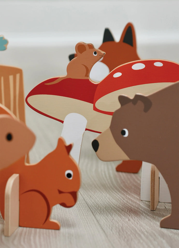 A squirrel, bear, and fox wooden toys gather around a red and white mushroom on a light wooden surface, resembling pieces from the Woodland Indoor Croquet Set.