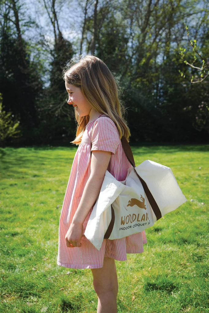 In a sunny garden, a young girl with long hair wears a pink striped dress and carries a white tote bag. Beside her, the Woodland Indoor Croquet Set rests on the vibrant grass among lush trees, inviting her to play in the enchanting forest glade.