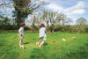 In a grassy field under a partly cloudy sky, two children play in a forest glade with toy wooden animals and a Woodland Indoor Croquet Set. One child grips a mallet-shaped stick, while the other concentrates on the scattered toys.