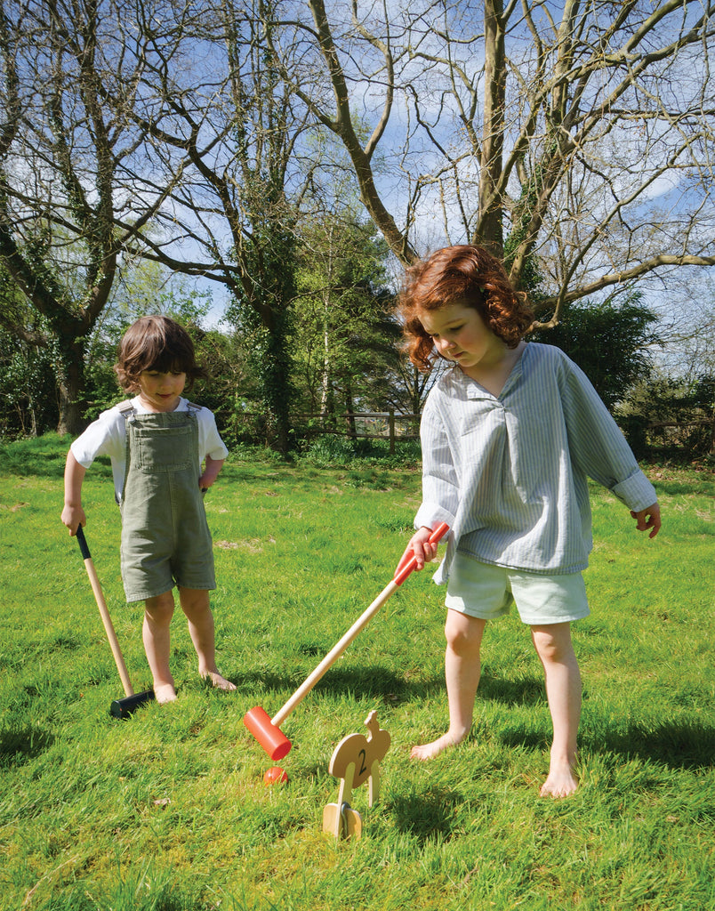 Two children play with a Woodland Indoor Croquet Set in a grassy field. The child on the left, wearing gray overalls, holds a mallet while the one on the right, in a striped shirt, is ready to hit a ball through an arch. Leafless trees frame their forest glade game.