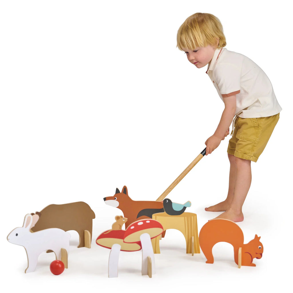 A young child in a white shirt and yellow shorts joyfully plays with the Woodland Indoor Croquet Set, using wooden mallets to interact with colorful animal and mushroom cutouts on the floor, featuring playful shapes like foxes, squirrels, and rabbits for imaginative adventures.