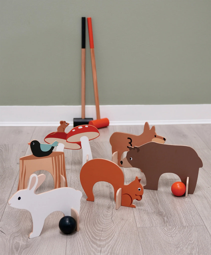 The Woodland Indoor Croquet Set features animal-shaped wooden arches, such as a rabbit, bear, and squirrel. The set includes red and black balls, with wooden mallets resting on a light wooden floor against a green wall, creating an inviting scene for imaginative forest glade play.