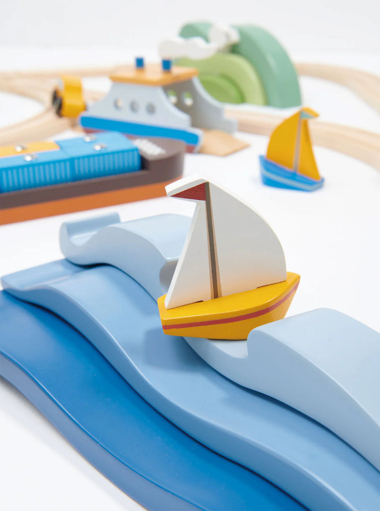 A close-up of a colorful children's wooden toy set featuring boats and waves. In the foreground, a small sailboat with a yellow and white design glides over blue solid wood waves. In the background, additional boats and sea creatures are partially visible, encouraging open-ended play with the Blue Water and Sail Boat Set.