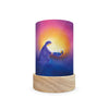 The Toverlux Lantern Shade Set - Holidays, featuring a cylindrical design with a wooden base, beautifully displays silhouettes of lanterns cradling a baby against a vibrant sunset. The enchanting shades seamlessly transition from deep purple and blue to bright orange and yellow, resulting in a captivating display.