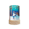 The Toverlux Lantern Shade - Seasons beautifully captures a winter scene with magical silhouettes of two figures in warm clothing holding hands. Snow-covered trees and glowing stars embellish the deep blue sky, creating an enchanting atmosphere. This charming piece rests on a wooden base, exuding warmth and wonder. (Ships in approximately one week.)