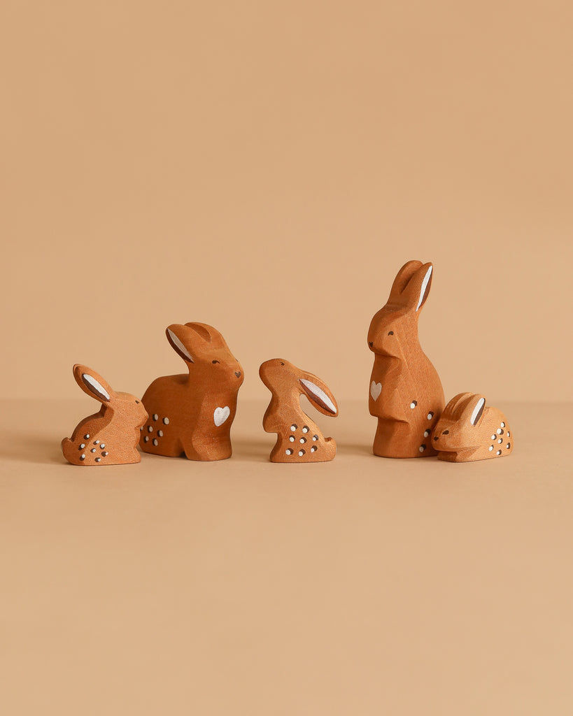 The Bumbu Handmade Wooden Brown Bunny Rabbit Family Set features five small, brown bunny figurines with white spots and heart details, arranged in a row on a beige background—ideal for Montessori toy collections.