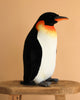 The Penguin Stuffed Animal, Medium, features lifelike black, white, and orange details and sits upright on a wooden stool against a beige background, showcasing the realistic design found in HANSA animals and similar plush toys.