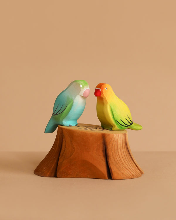 Two handmade wooden bird figurines—one blue-green, one yellow-orange—from the Bumbu Handmade Wooden Tree Stump and Lovebirds Set perch closely on a carved tree stump, set against a plain beige background.
