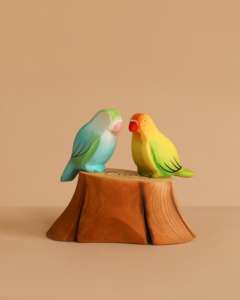 Two handmade wooden bird figurines—one blue-green, one yellow-orange—from the Bumbu Handmade Wooden Tree Stump and Lovebirds Set perch closely on a carved tree stump, set against a plain beige background.