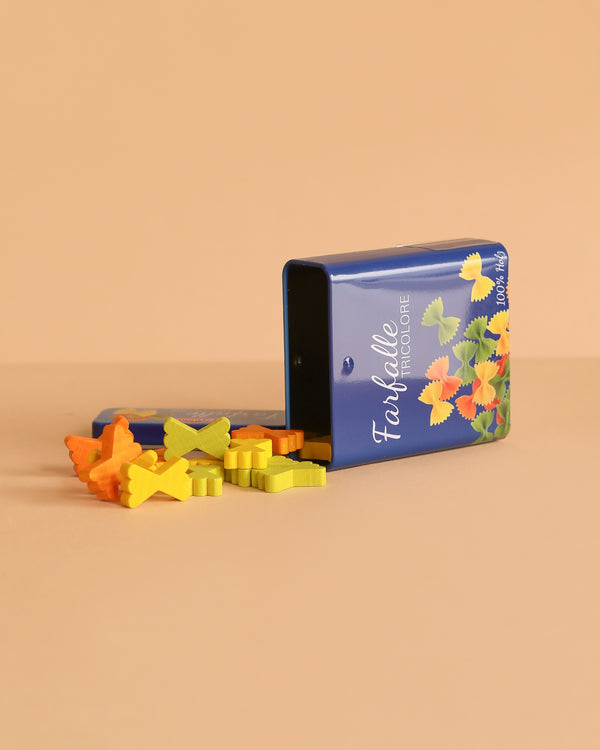 A blue tin labeled "Erzi Farfalle Pasta Pretend Food" lies sideways, spilling wooden toy bowtie pasta across a peach surface and resembling miniature food.