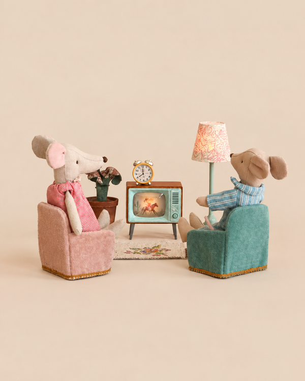 Two Maileg mice sit in miniature armchairs facing a small vintage-style television set, an alarm clock, and a floor lamp in this Maileg Movie Night Set.